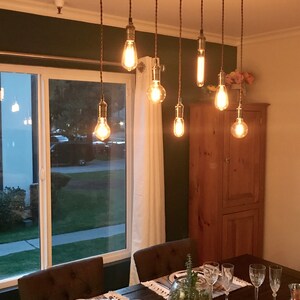May include: A rustic wooden dining room chandelier with eight Edison bulb lights hanging from rope cords. The chandelier is over a dark wood dining table set with white placemats, glasses, and a potted succulent.