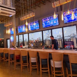 May include: A long wooden bar with a row of bar stools. The bar is in a brewery with large stainless steel tanks and televisions playing sports. The sign behind the bar says "Sandra for GJ".