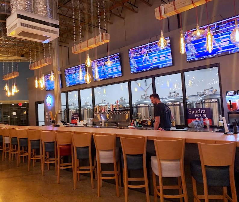 May include: A long wooden bar with a row of bar stools. The bar is in a brewery with large stainless steel tanks and televisions playing sports. The sign behind the bar says "Sandra for GJ".