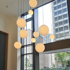 May include: A modern interior features a cluster of spherical pendant lights in varying sizes. The globes are a soft, warm white, suspended from black cords against a backdrop of large windows and city buildings.