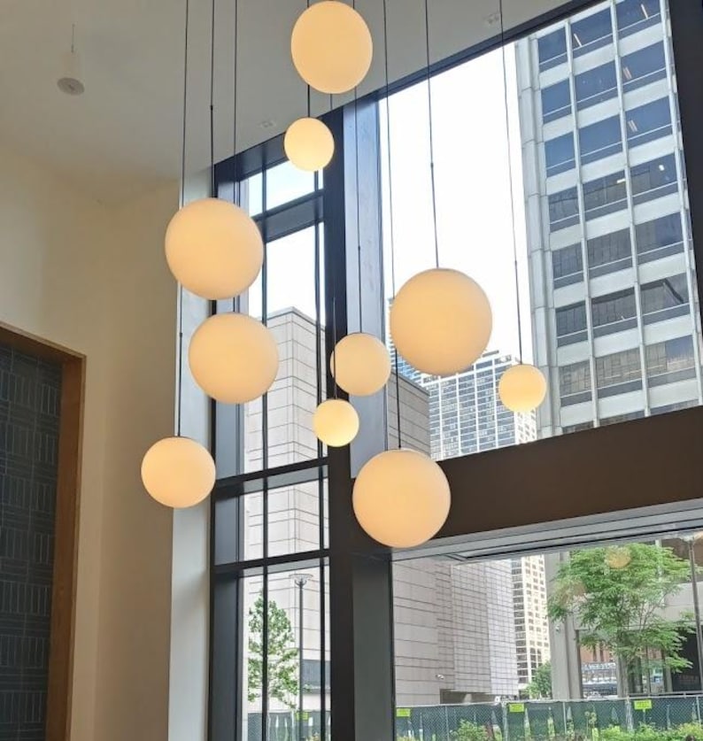 May include: A modern interior features a cluster of spherical pendant lights in varying sizes. The globes are a soft, warm white, suspended from black cords against a backdrop of large windows and city buildings.