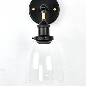 May include: Black wall sconce with a clear glass shade. The sconce has a gold adjustable arm and a black metal base.