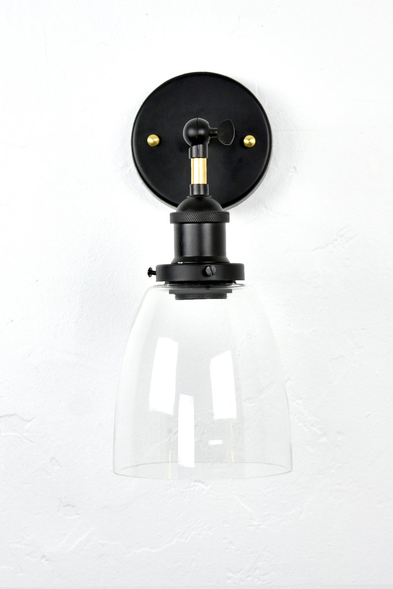 May include: Black wall sconce with a clear glass shade. The sconce has a gold adjustable arm and a black metal base.