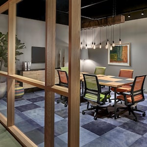 May include: A modern office meeting room with a wooden table and chairs. The room has a checkered blue and grey carpet, a large window, and a rustic wooden beam with hanging light fixtures.