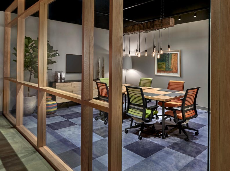 May include: A modern office meeting room with a wooden table and chairs. The room has a checkered blue and grey carpet, a large window, and a rustic wooden beam with hanging light fixtures.