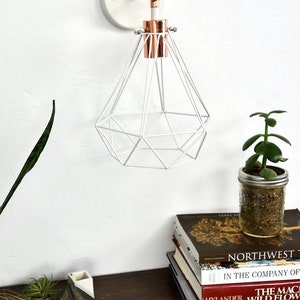 May include: White geometric pendant light with copper accents, mounted on a white wall. A small potted plant in a mason jar sits on a wooden surface, along with a stack of books. The books include titles like "Northwest" and "Wild Flowers".