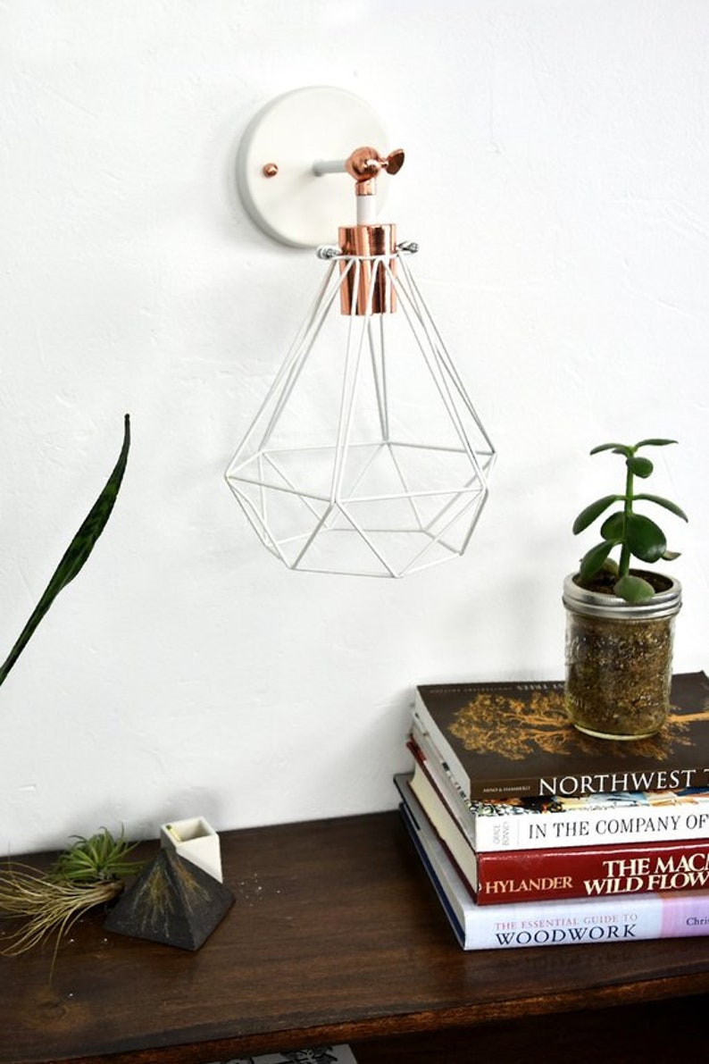 May include: White geometric pendant light with copper accents, mounted on a white wall. A small potted plant in a mason jar sits on a wooden surface, along with a stack of books. The books include titles like "Northwest" and "Wild Flowers".