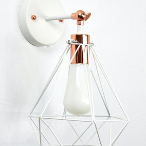 May include: White and copper-colored wall sconce with a geometric cage design. The light fixture features a white bulb and a copper-colored accent around the bulb base. The sconce is mounted on a white circular base.