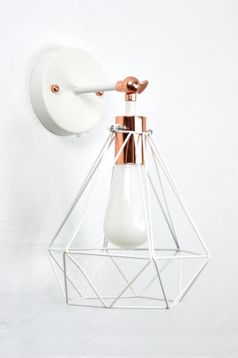 May include: White and copper-colored wall sconce with a geometric cage design. The light fixture features a white bulb and a copper-colored accent around the bulb base. The sconce is mounted on a white circular base.
