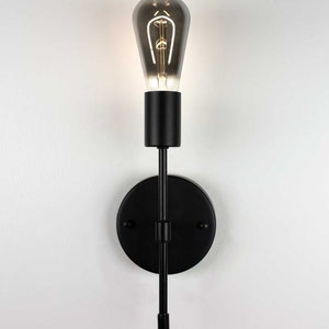 K&ouml;nnte beinhalten: Schwarze Wandleuchte mit einer einzelnen Gl&uuml;hbirne. Die Leuchte hat einen runden Sockel und einen langen, geraden Arm, der sich nach oben erstreckt. Die Gl&uuml;hbirne ist eine klare Edison-Gl&uuml;hbirne aus Glas mit mattem Finish.
