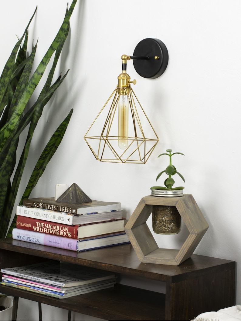 May include: A gold geometric wall sconce with a black base and a vintage-style lightbulb. A small wooden hexagonal planter holds a glass jar with a plant. Books and magazines are stacked on a dark wooden shelf.