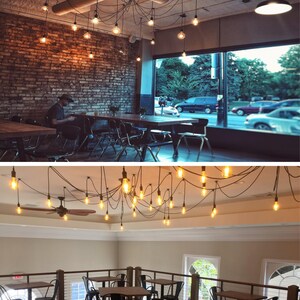 May include: A cafe interior with exposed brick walls and a large window. The cafe has multiple tables and chairs, and a unique light fixture with many Edison bulbs hanging from the ceiling.