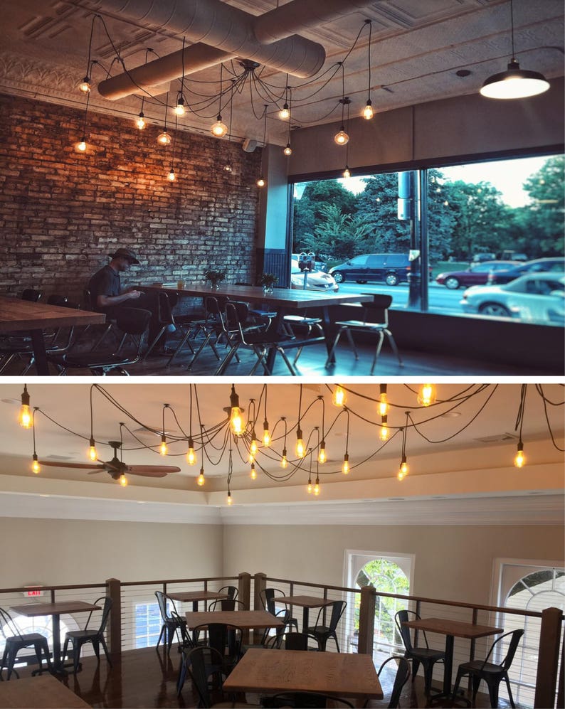 May include: A cafe interior with exposed brick walls and a large window. The cafe has multiple tables and chairs, and a unique light fixture with many Edison bulbs hanging from the ceiling.