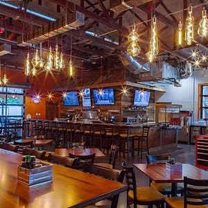 May include: A large, open-plan restaurant with wooden tables and chairs. The restaurant has a bar with a wooden counter and stools. There are multiple large televisions mounted on the wall behind the bar. The restaurant has a rustic, industrial feel with exposed beams and ductwork. There are many Edison bulb light fixtures hanging from the ceiling.