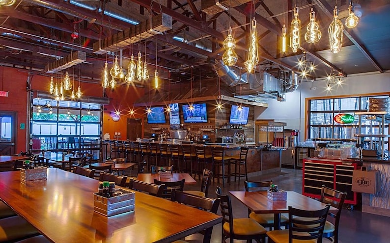 May include: A large, open-plan restaurant with wooden tables and chairs. The restaurant has a bar with a wooden counter and stools. There are multiple large televisions mounted on the wall behind the bar. The restaurant has a rustic, industrial feel with exposed beams and ductwork. There are many Edison bulb light fixtures hanging from the ceiling.