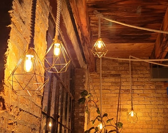 May include: A brick wall with a series of gold cage light fixtures hanging from the ceiling. The lights are connected by white rope and are lit with warm white bulbs.