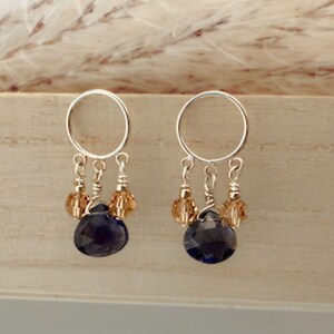 Peut inclure: Boucles d'oreilles en argent avec un cercle en haut, des brins de petites perles ambrées et une grosse pierre en forme de goutte d'eau bleu foncé. Les boucles d'oreilles sont présentées sur une surface en bois clair.