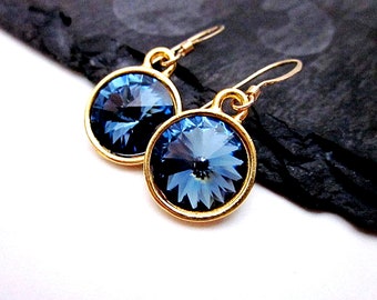 May include: A pair of gold-toned drop earrings featuring large, round, faceted blue crystals. The crystals are set in a gold bezel, and the earrings have a simple hook closure. The earrings are displayed on a dark, textured surface.