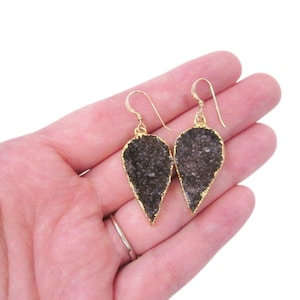 Può includere: Un paio di orecchini a goccia con un centro grigio scuro strutturato e un bordo color oro. Gli orecchini pendono da ganci color oro. Gli orecchini sono esposti su uno sfondo chiaro.