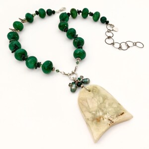 May include: A necklace featuring faceted green beads strung along a silver-toned chain, accented with smaller dark green beads and a central silver-toned horseshoe charm. A large, flat, cream-colored stone pendant hangs from the horseshoe charm.