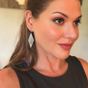 May include: A silver-toned dangle earring featuring a diamond-shaped pendant with an intricate, openwork lattice pattern. The earring hangs from a thin metal hook, showcasing a geometric design that creates a delicate, airy appearance.