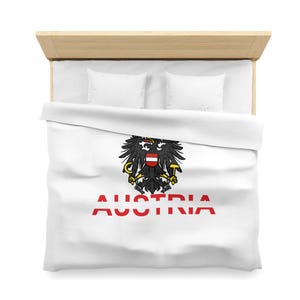 Puede incluir: Funda nórdica blanca con el escudo de armas austriaco, un águila negra con un escudo rojo y blanco, y la palabra "AUSTRIA" en rojo. La ropa de cama está sobre una cama de madera clara.