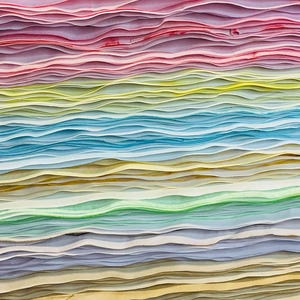 May include: A stack of thin, wavy paper sheets arranged in a horizontal gradient pattern. The colors transition from deep pink and red at the top, through yellow, light blue, and green, to soft purple and beige tones at the bottom of the stack.