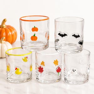 May include: Five clear glass tumblers with colourful rims and small raised motifs. One has orange pumpkins, one has black bats, one has yellow rubber ducks, one has red and orange autumn leaves, and one has white ghost figures.
