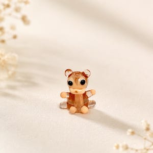 May include: A small, translucent brown glass figurine shaped like a bear. The bear has two round ears, two black dot eyes, and light peach-colored accents on its snout, hands, and feet. The glass has a smooth, glossy finish throughout the piece.