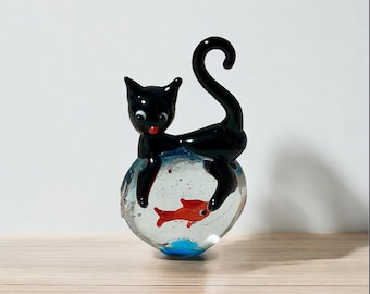 Può includere: Una statuetta in vetro raffigurante un gatto nero lucido con la coda arricciata, appollaiato sopra una ciotola di vetro trasparente e arrotondata. All'interno della ciotola è visibile un piccolo pesce color arancione vivo, occhi bianchi e naso rosso.