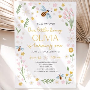 May include: A bee-themed first birthday party invitation with a floral border. The invitation says "Buzz on over, Our little honey Olivia is turning one. Join us to celebrate! June 1st, 11:00 am. The Anderson Residence, 123 Daisy Street, New York, NY. RSVP by March 15th to Elizabeth at +123 456 789."