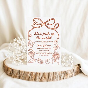 May include: A red and white bridal shower invitation with a hand-drawn illustration of a bow and various fruits and vegetables. The text reads "She's fresh off the market" and includes details about the event, including the date, time, location, and RSVP information.