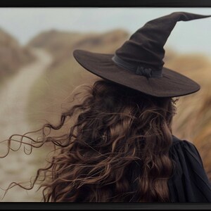 May include: A framed print featuring a person wearing a black witch hat, facing away from the viewer. The person has long, wavy brown hair. The background shows a path through tall, dry grass.
