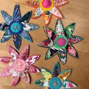 May include: Five handmade fabric flower decorations with colorful patchwork petals and felt centers. Each flower has a unique color scheme, including pink, blue, orange, and green. The flowers are arranged on a wooden surface.