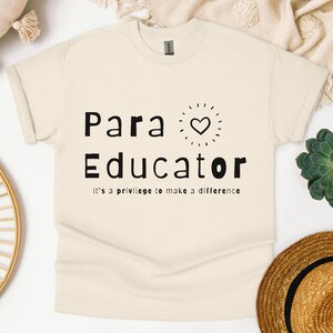 Może przedstawiać: Beżowa koszulka z czarnym napisem "Para Educator it's a privilege to make a difference". Małe serce z promieniami słońca wokół znajduje się nad słowem "Educator".