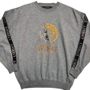 May include: A gray sweatshirt with a large embroidered design featuring a face with gold and black details. The sleeves have black stripes with the text "A.VERSACE" in gold. The word "A.VERSACE" is also embroidered in gold below the face design.