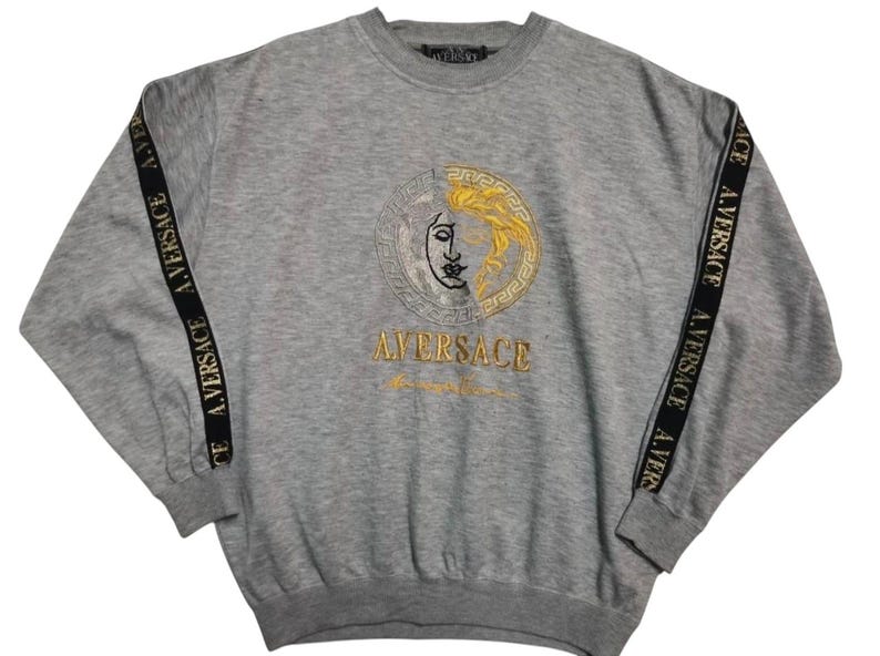 May include: A gray sweatshirt with a large embroidered design featuring a face with gold and black details. The sleeves have black stripes with the text "A.VERSACE" in gold. The word "A.VERSACE" is also embroidered in gold below the face design.