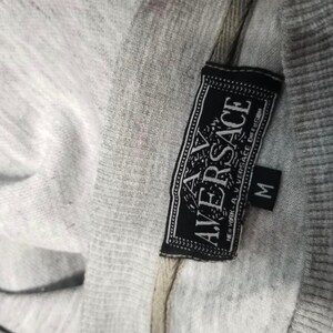 May include: Close-up of a gray sweatshirt with a black label. The label reads "AV VERSACE" in white lettering, with a smaller "M" size tag. The fabric appears to be a soft, knit material.