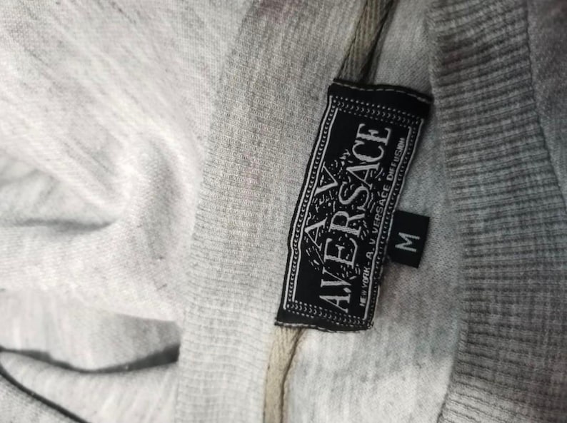 May include: Close-up of a gray sweatshirt with a black label. The label reads "AV VERSACE" in white lettering, with a smaller "M" size tag. The fabric appears to be a soft, knit material.