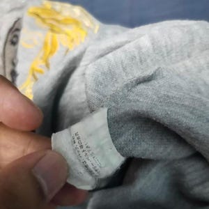 May include: Close-up of a gray t-shirt with a tag. The tag has Japanese text. The shirt has a yellow design on the upper left side. The shirt is folded.