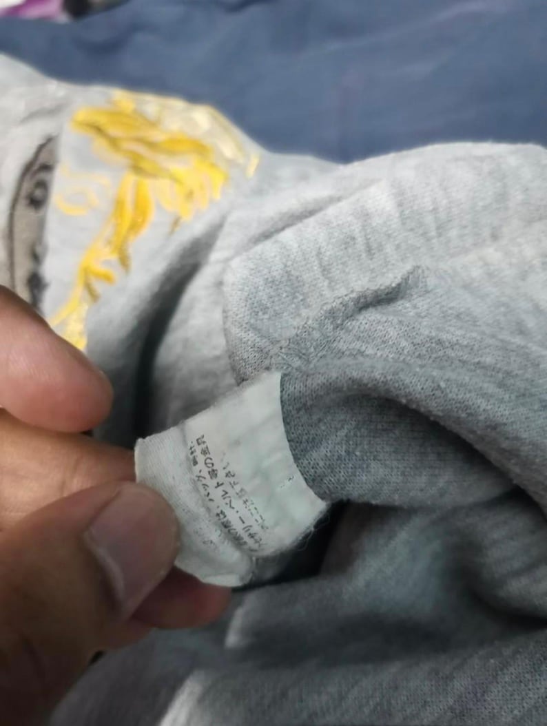 May include: Close-up of a gray t-shirt with a tag. The tag has Japanese text. The shirt has a yellow design on the upper left side. The shirt is folded.