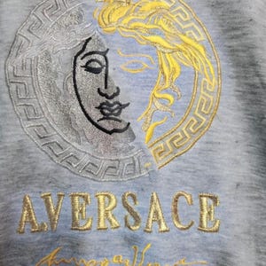 May include: A light blue sweatshirt featuring an embroidered design of a split Medusa head in silver and gold. The word "AVERSACE" is embroidered in gold, with a gold signature below the word.