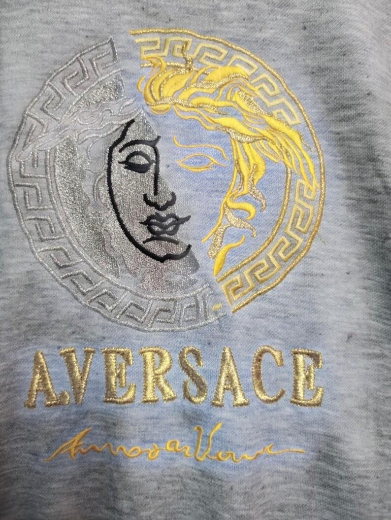 May include: A light blue sweatshirt featuring an embroidered design of a split Medusa head in silver and gold. The word "AVERSACE" is embroidered in gold, with a gold signature below the word.