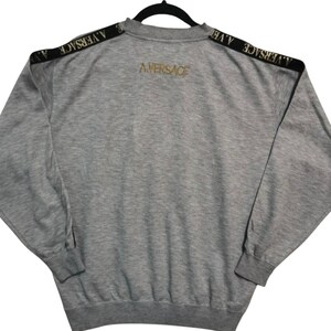 May include: A gray sweatshirt with black shoulder stripes featuring the text "A. VERSACE". The back of the sweatshirt has the text "A. VERSACE" in gold lettering. The sweatshirt is hanging on a black hanger.