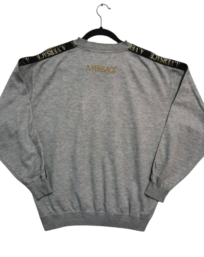 May include: A gray sweatshirt with black shoulder stripes featuring the text "A. VERSACE". The back of the sweatshirt has the text "A. VERSACE" in gold lettering. The sweatshirt is hanging on a black hanger.