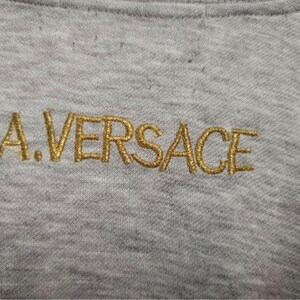 May include: Close-up of a gray fabric with the embroidered text "A.VERSACE" in gold thread. The letters are stylized and appear on the upper portion of the fabric, suggesting a garment detail.