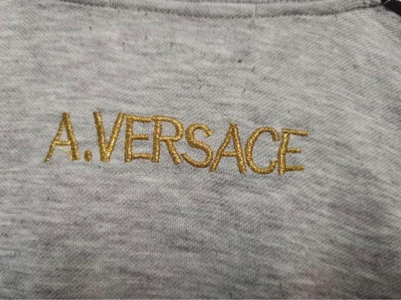 May include: Close-up of a gray fabric with the embroidered text "A.VERSACE" in gold thread. The letters are stylized and appear on the upper portion of the fabric, suggesting a garment detail.