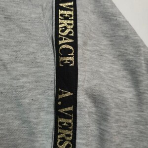 May include: Close-up of a gray sweatshirt with a black vertical stripe. The stripe features the gold text "A. VERSACE" repeated down its length. The fabric has a textured appearance.