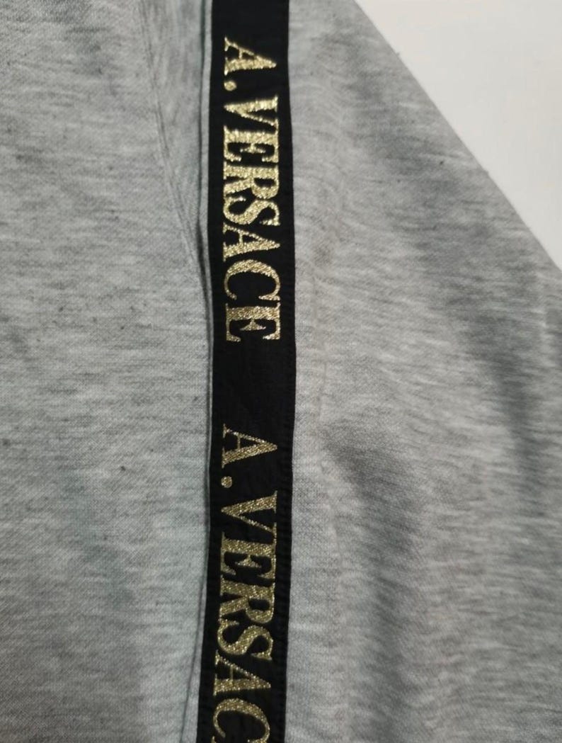 May include: Close-up of a gray sweatshirt with a black vertical stripe. The stripe features the gold text "A. VERSACE" repeated down its length. The fabric has a textured appearance.