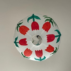 May include: A round, white paper lantern decorated with hand-painted red and orange tulips and green leaves. The floral design is arranged around the lantern's circumference. The lantern is suspended from the ceiling.